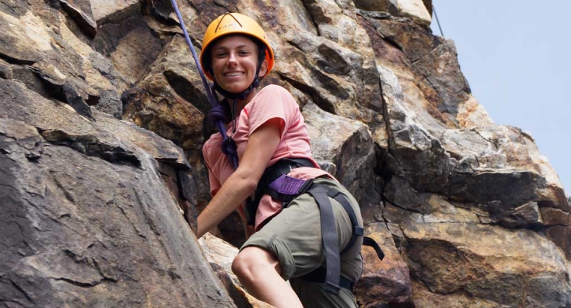 A person wearing safety gear is secured by ropes as they pause climbing a rock wall to smile for the photo.